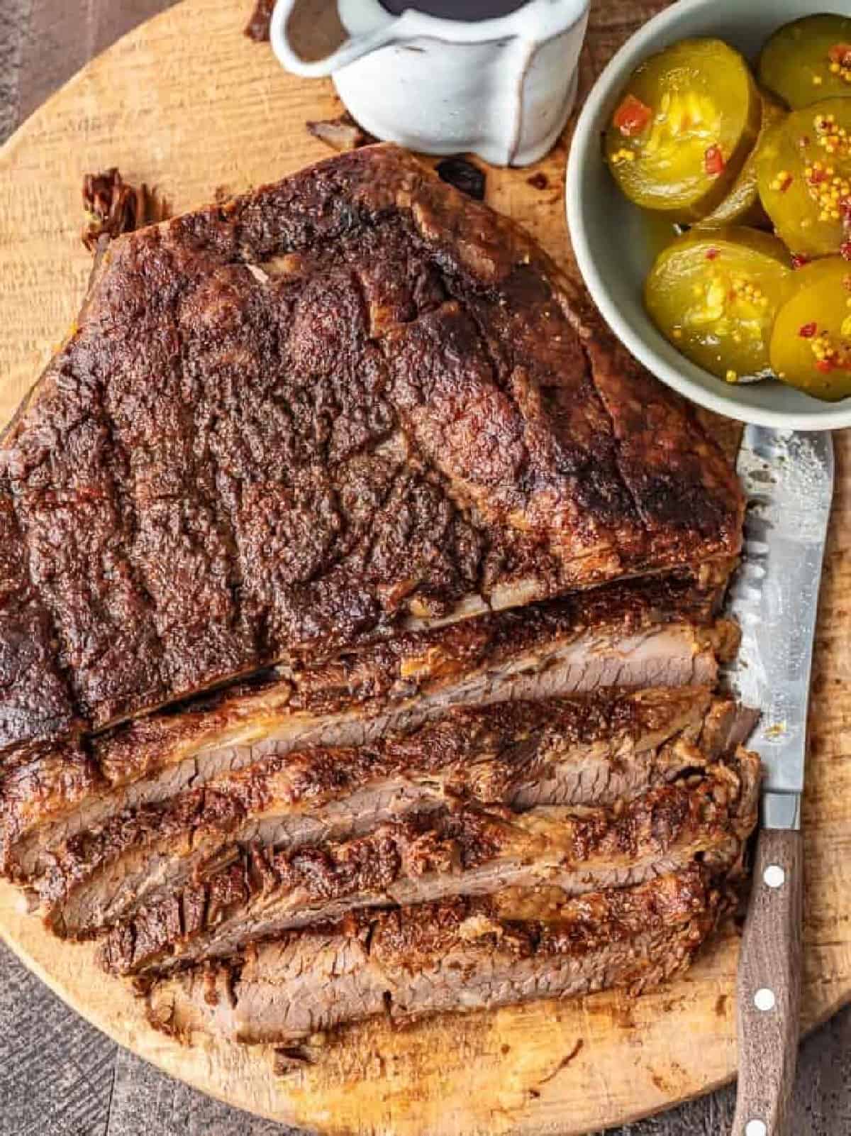sliced brisket on cutting board with a knife and a bowl of pickles.